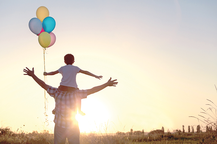 Zu sehen ist ein Mann der ein Kind auf den Schultern trägt. Beide breiten ihre Arme zur Seite aus und gucken in die Sonne. Das Kind hält mehrere bunte Luftballons in der Hand. Sie stehen auf einem Feld.